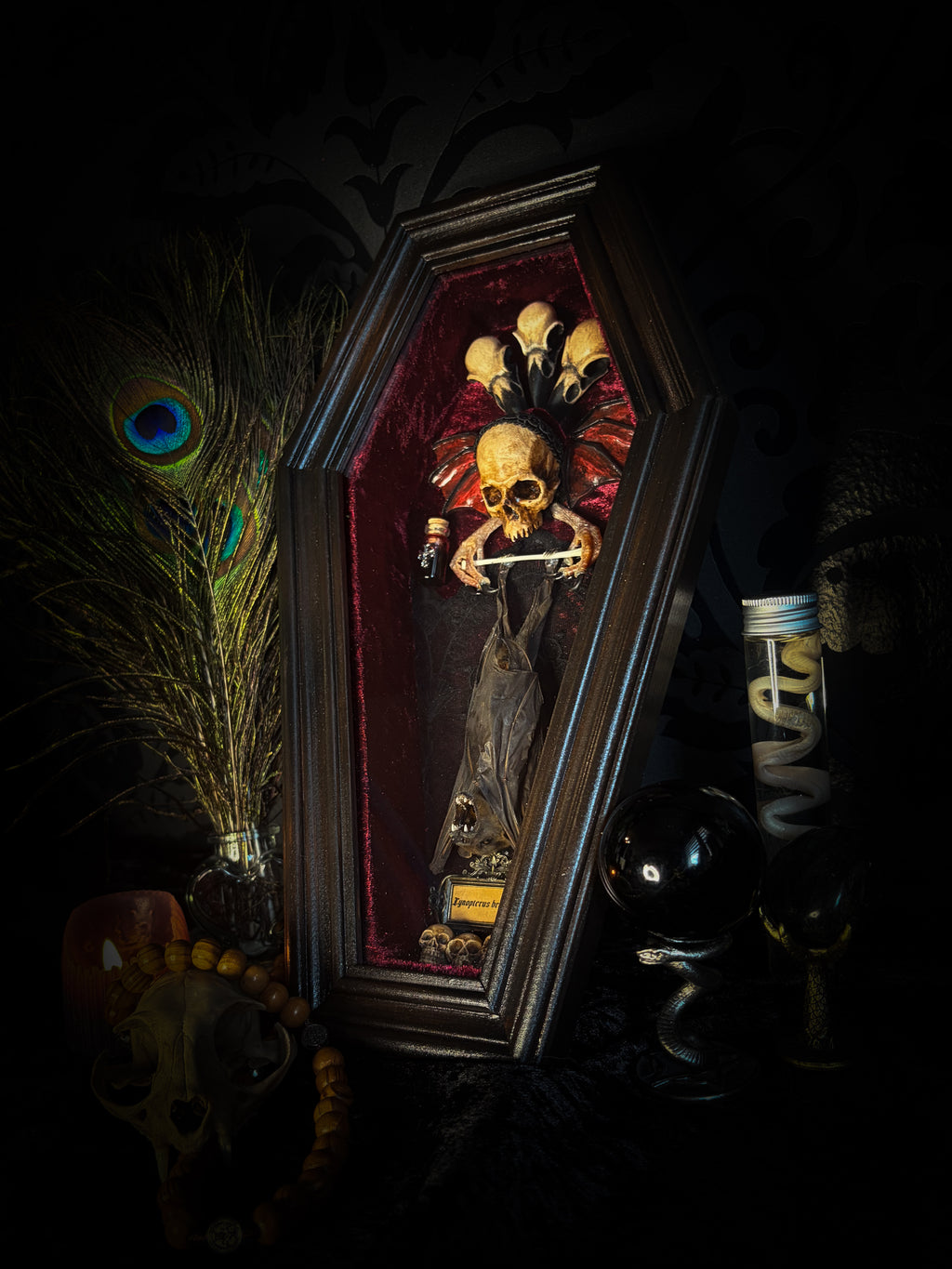 Sanguis Aeternus: mummified bat in a handmade wooden coffin with burgundy velvet, a hooded winged vampire skull, raven skull ornaments and decorative blood vials.

