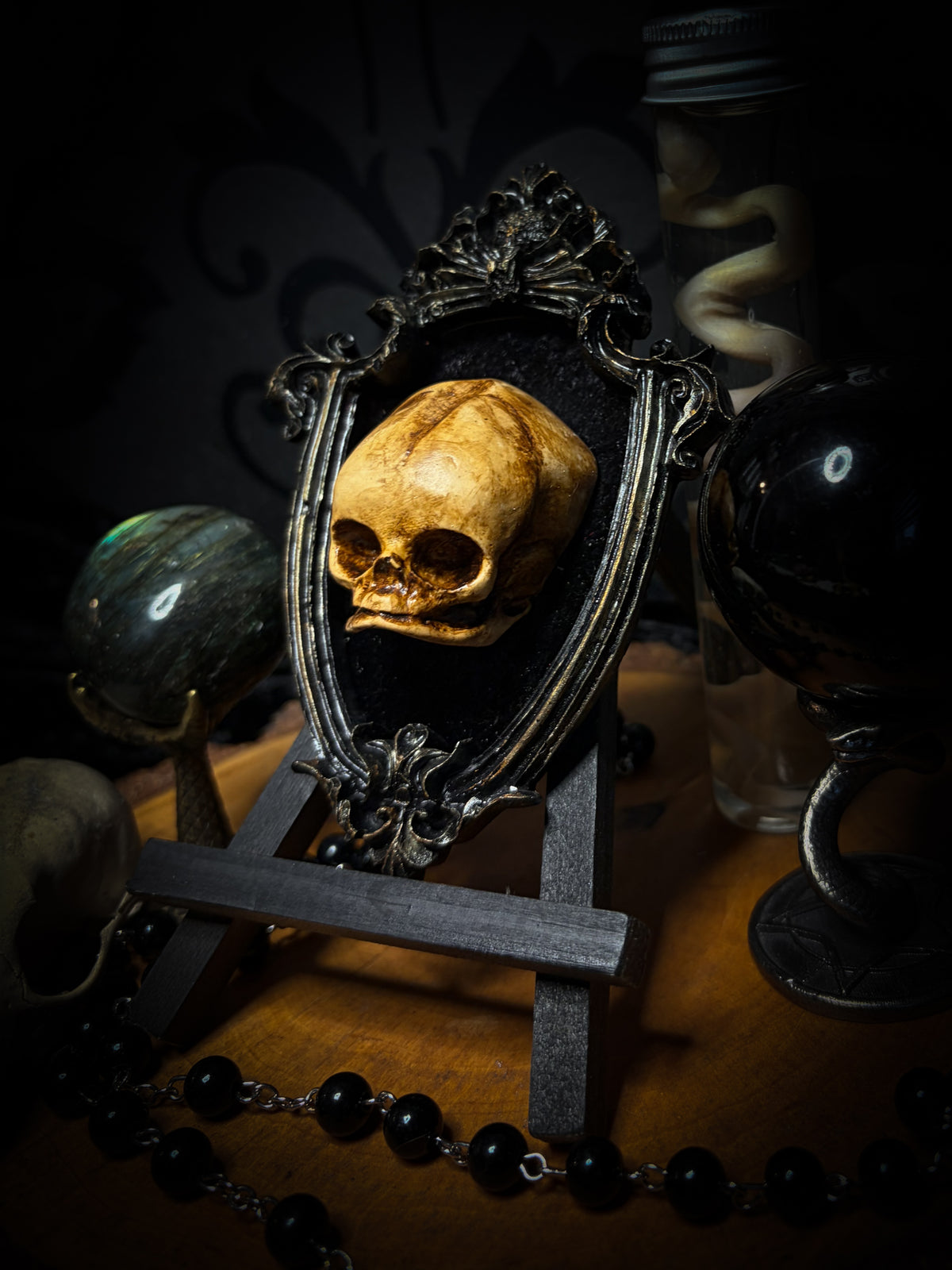 Little Vigil by Morbid Oddities, a patinated skull cast on black velvet in an ornate dark shield-shaped frame with aged metallic detailing.⠋

