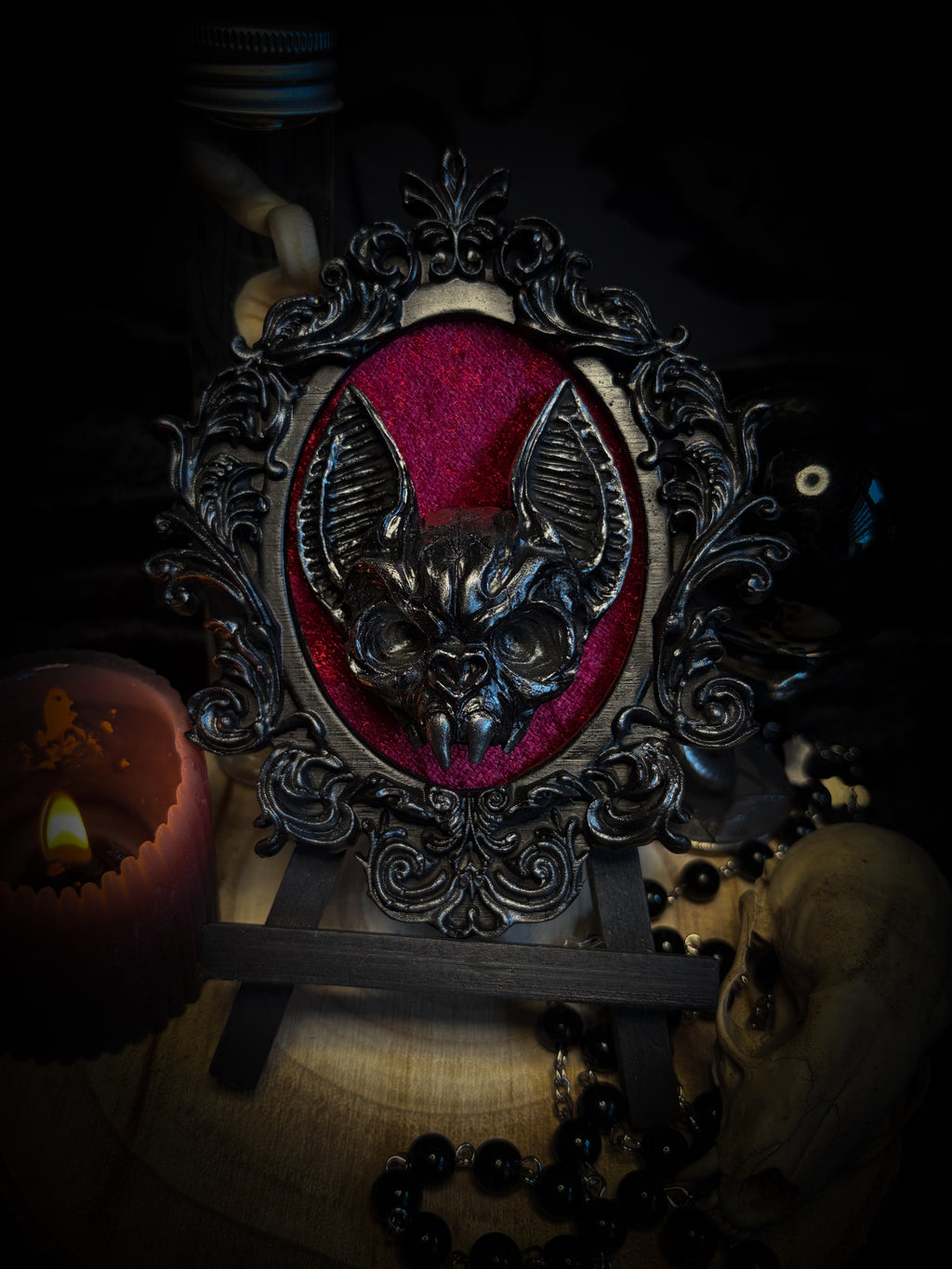 Decorative still life with a candle, skull, and framed picture on a dark background Handmade gothic wall frame featuring a detailed bat centerpiece set on deep blood-red velvet, enclosed in an ornate baroque-style frame. Dramatic, ritual-inspired made-to-order piece with rich crimson velvet and dark gothic aesthetic, 12.5x10cm | Morbid Oddities